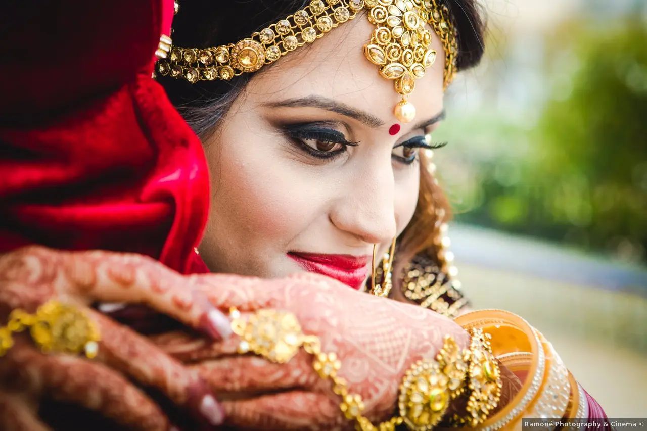 Una novia con joyas de oro, lápiz labial rojo y un tocado adornado abraza a alguien, con las manos visibles.