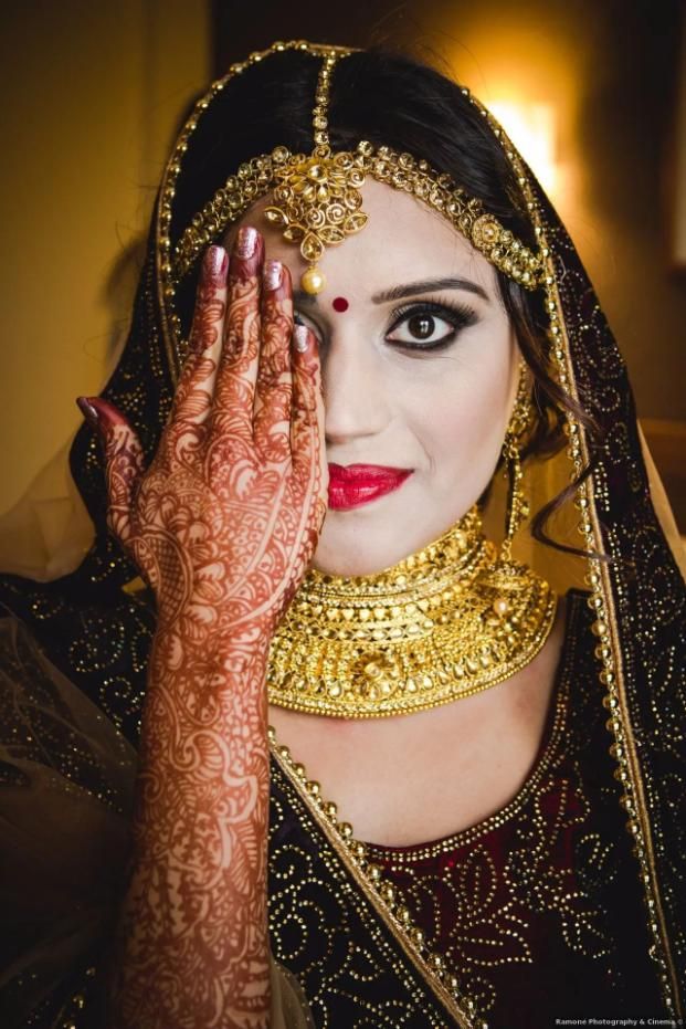 Mujer con mano pintada de henna cubriendo media cara, adornada con joyas de oro; labios rojos.