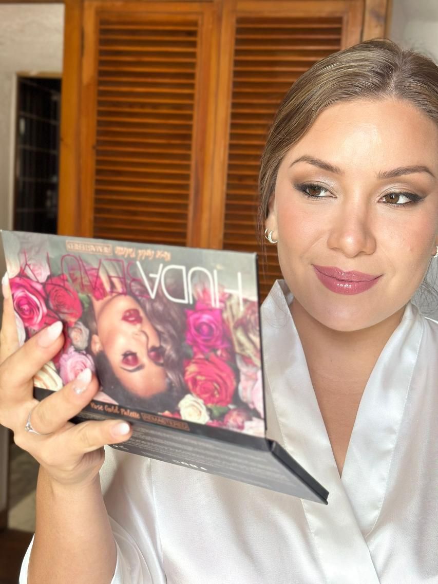 Mujer sosteniendo una paleta de sombras de ojos de Huda Beauty, vestida con una bata blanca, sonriendo.