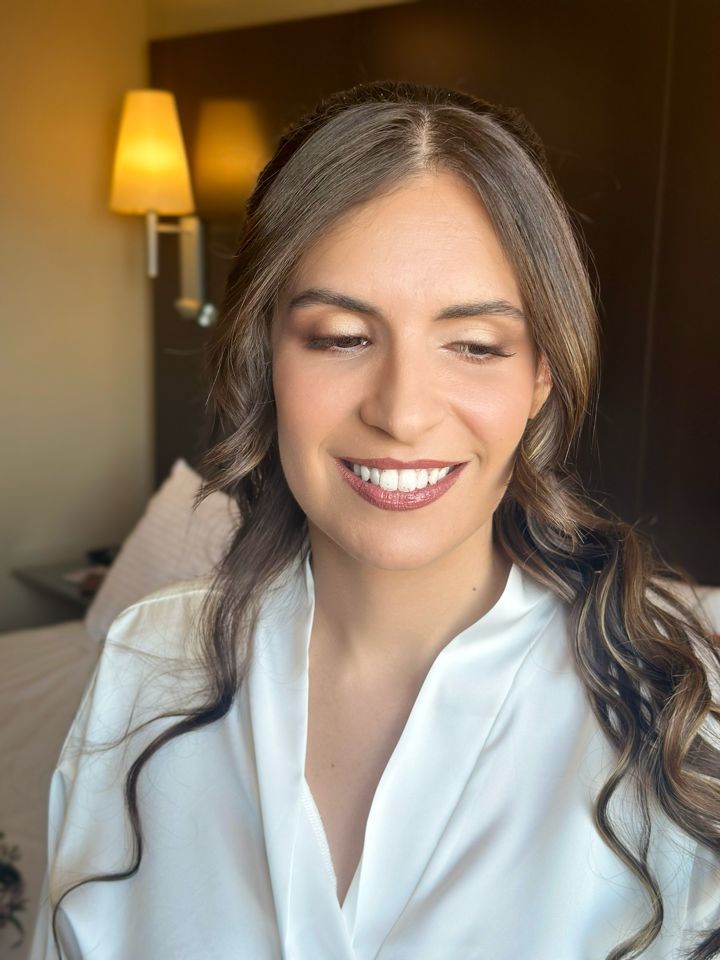 Mujer con cabello castaño ondulado, maquillada, sonriente, vistiendo una bata blanca, frente a la ventana de una habitación