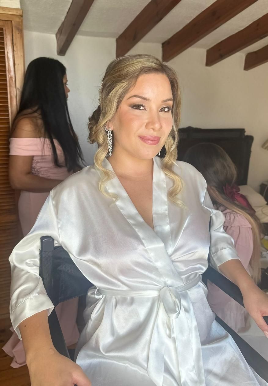 Mujer con túnica blanca, peinado recogido y pendientes, preparándose para un evento.