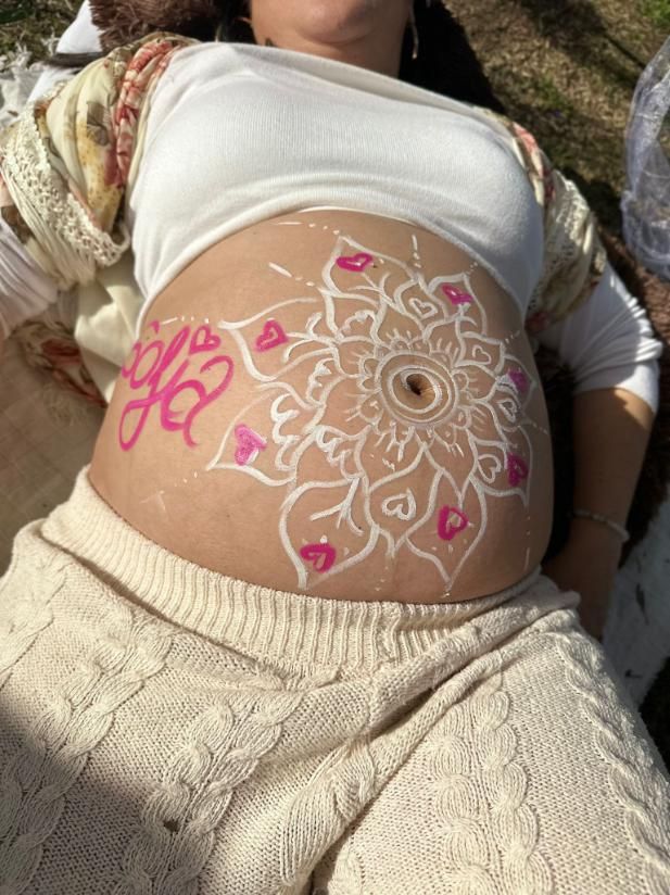 Vientre de embarazada pintado con un diseño floral blanco, corazones rosas y la palabra 