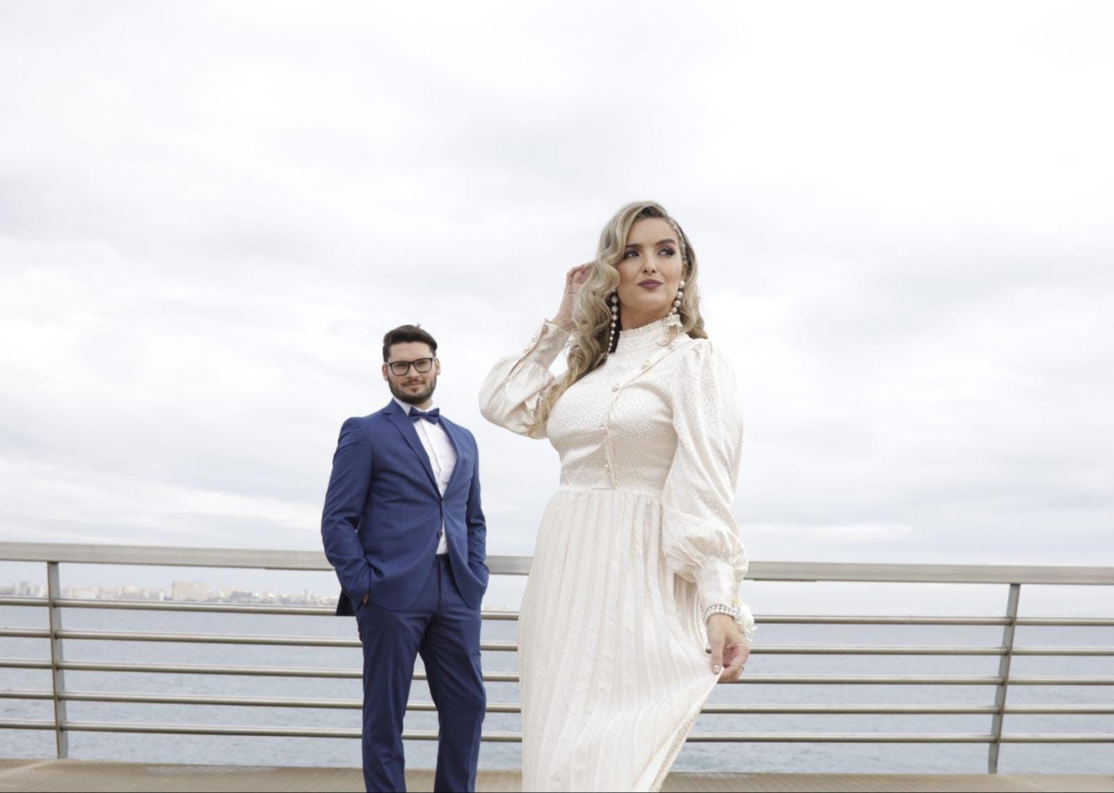 Pareja posando al aire libre cerca del agua; mujer con vestido blanco, hombre con traje azul.