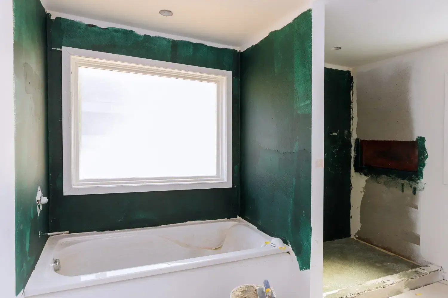 Renovación del baño en proceso; bañera y ventana enmarcadas por paredes pintadas de verde, puerta a la derecha.