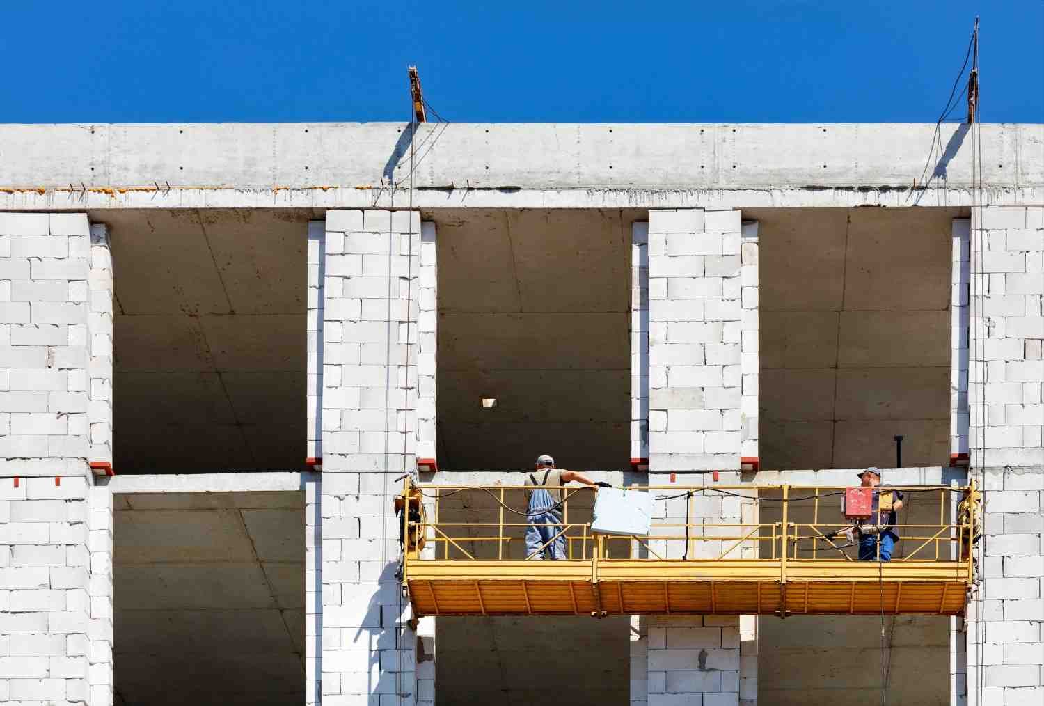 Obreros de la construcción en un andamio amarillo trabajando en un edificio blanco con ventanas abiertas. Cielo azul.