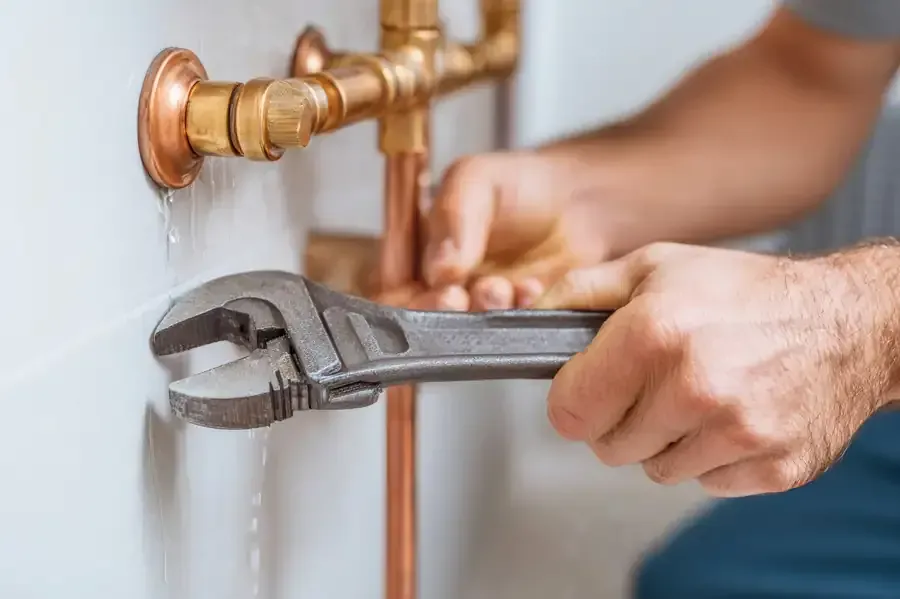 Plomero usando una llave inglesa sobre tuberías de cobre con fugas contra una pared blanca.