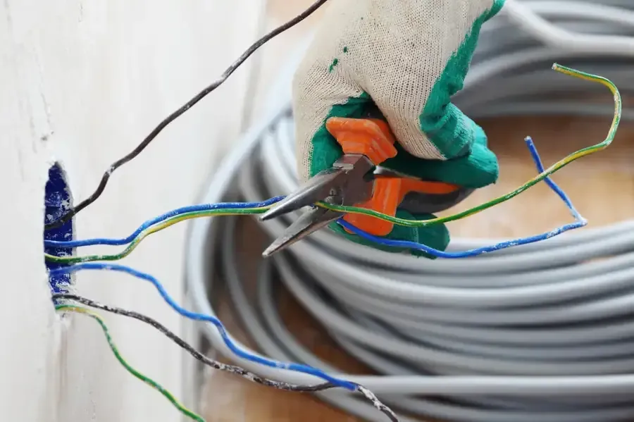 Mano enguantada que utiliza alicates para cortar cables que salen de una toma de corriente en una pared.