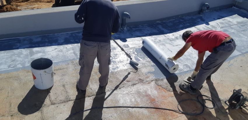 Dos trabajadores aplican un sellador blanco a una superficie de hormigón, utilizando rodillos y un material enrollado.