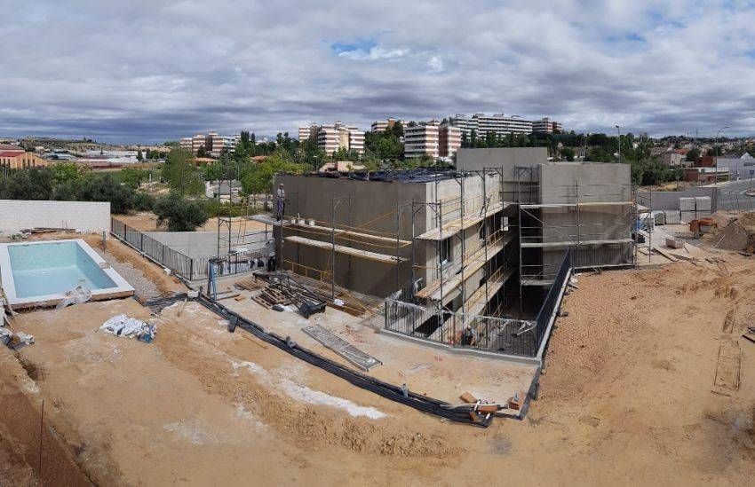 Sitio de construcción con edificio parcialmente construido, piscina y paisaje circundante bajo cielo nublado.