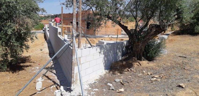 Sitio de construcción con un muro parcialmente construido, un árbol y una persona con una camisa roja.
