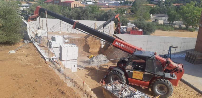 Un manipulador telescópico rojo levanta bloques en una obra. Un brazo de pluma negro se extiende sobre una zanja de tierra.