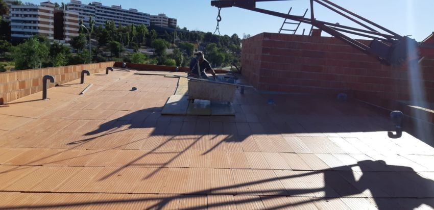 Obrero en un tejado. Pila de madera y grúa. Edificios y árboles al fondo. Luz solar intensa.