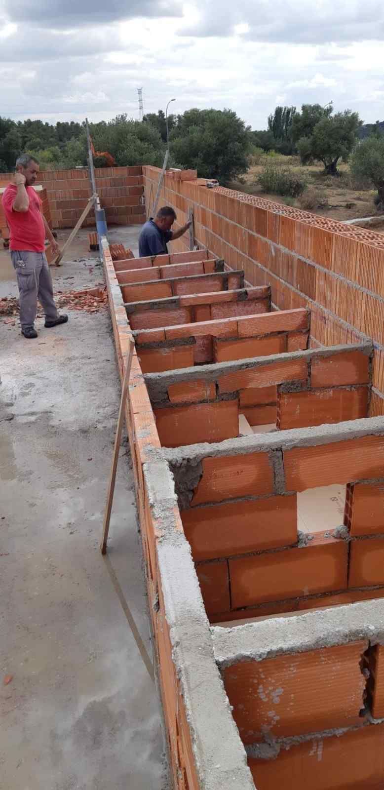 Obreros construyen una estructura de ladrillo al aire libre. Una persona con camisa roja trabaja. Cielo nublado.