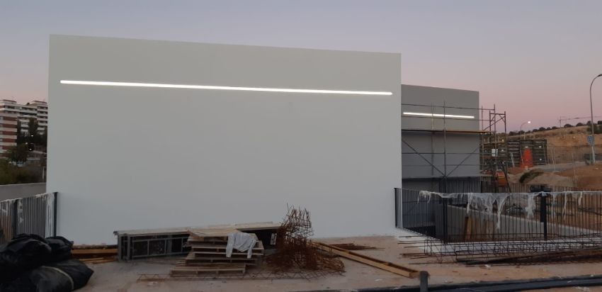 Vista exterior de un edificio blanco con una barra de luz horizontal, en construcción. Andamios visibles.