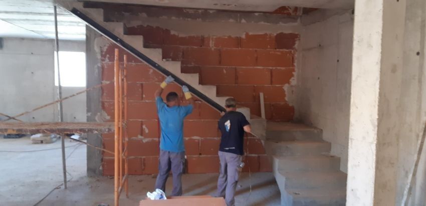 Dos obreros de la construcción cargan una viga larga por una escalera. Llevan ropa de trabajo y guantes.