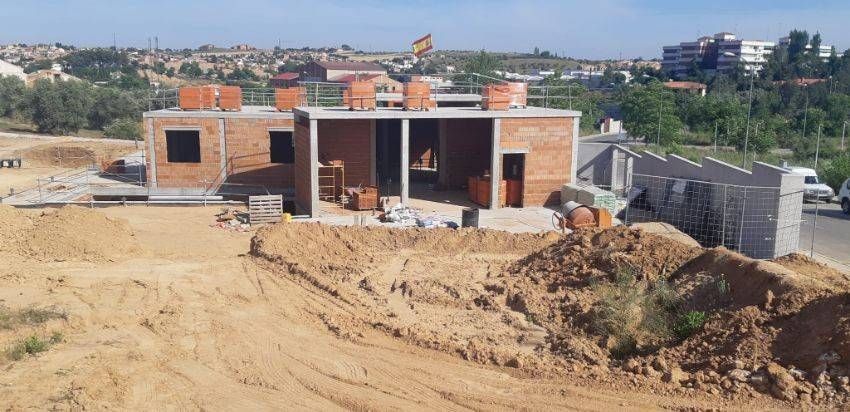 Un sitio de construcción con estructuras de ladrillo, montículos de tierra y un fondo suburbano bajo un cielo soleado.
