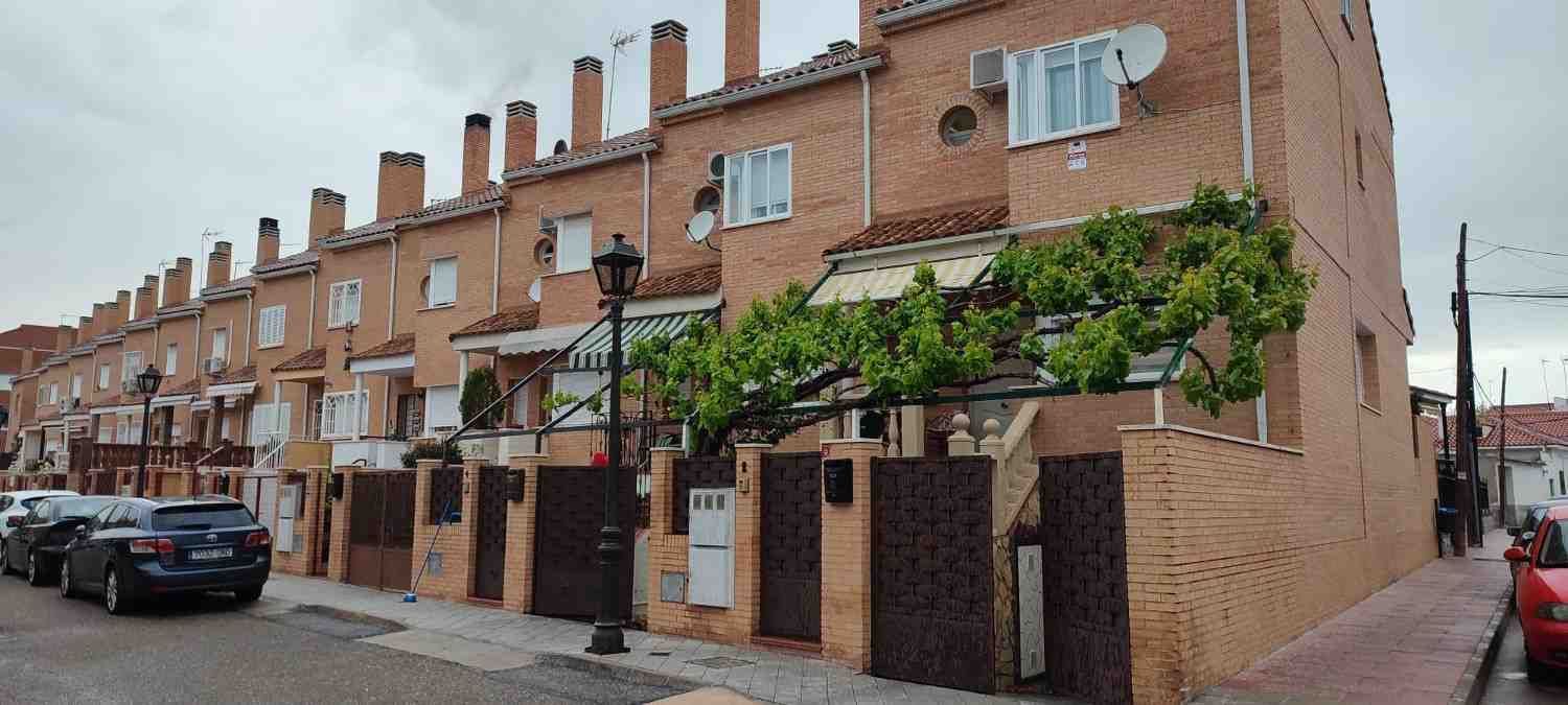Hilera de casas adosadas de ladrillo con chimeneas, portones oscuros y una calle. Día nublado.