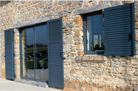 Extérieur d'un bâtiment en pierre avec des volets bleus sur une porte coulissante en verre et une fenêtre.