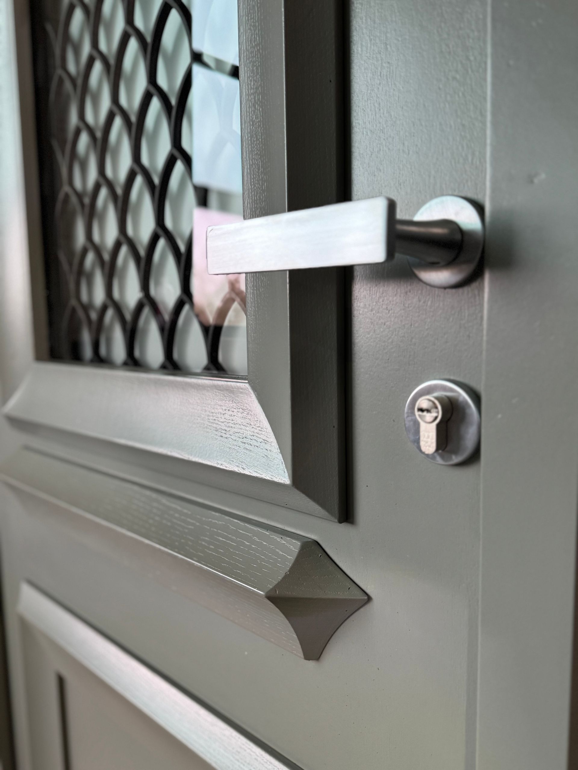 Porte grise avec poignée en métal, serrure et fenêtre grillagée décorative.