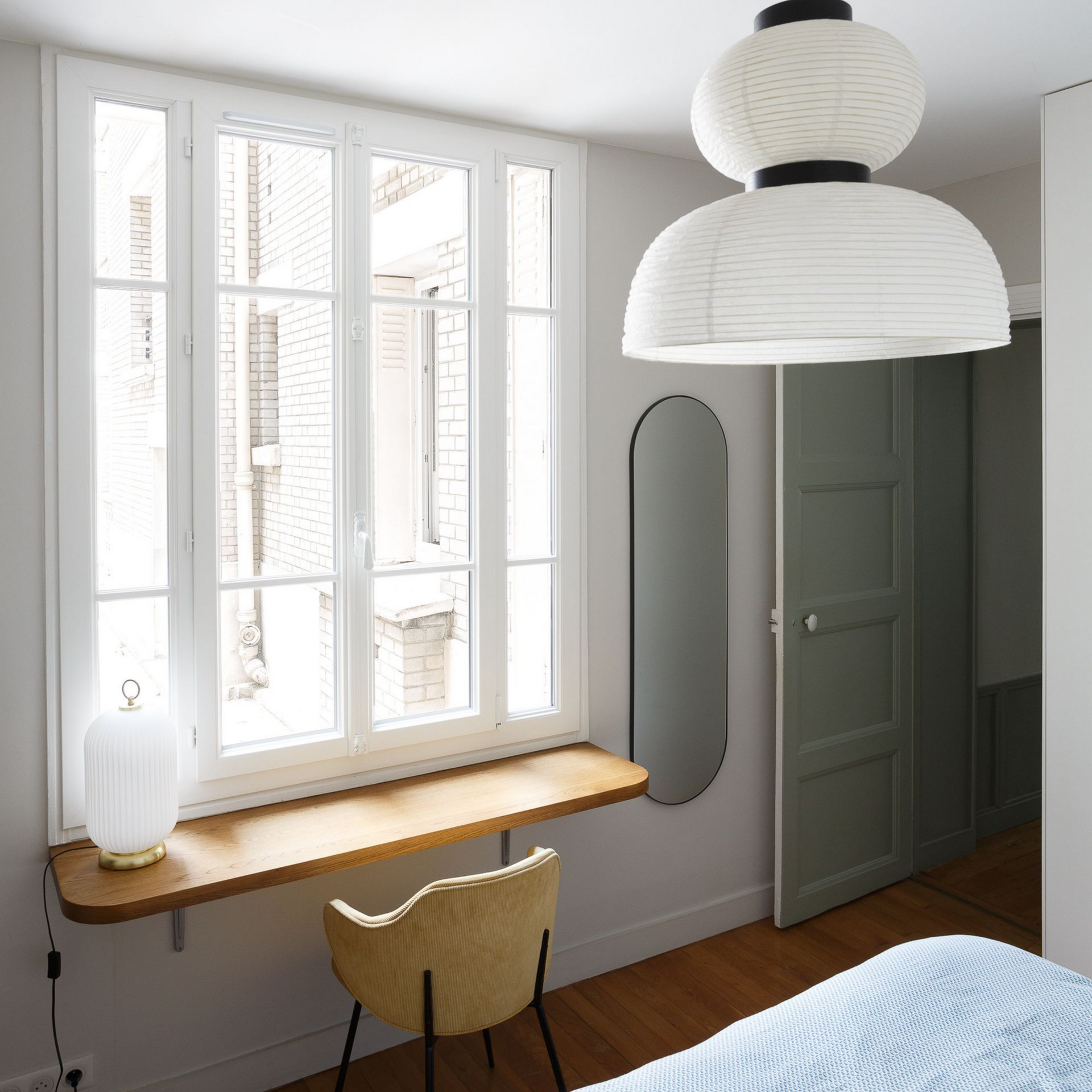 Chambre avec fenêtre, bureau, chaise et suspension ; porte à droite.