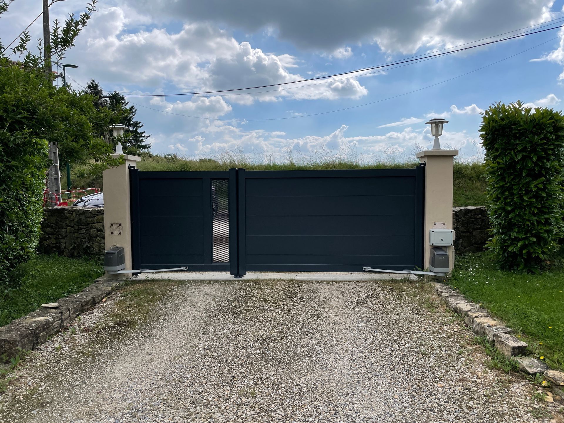 Portail d'allée gris foncé, piliers beiges, allée de gravier, ciel couvert.