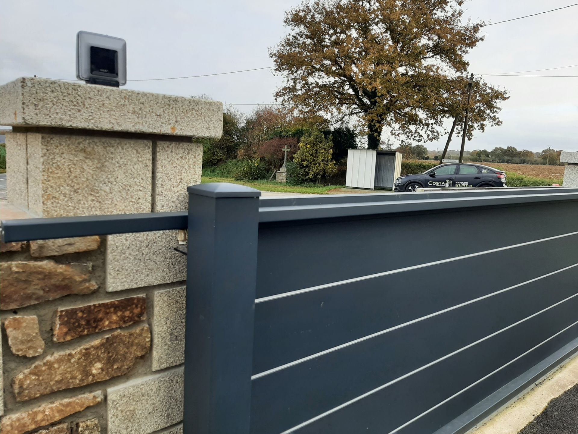 Portail coulissant gris à bandes blanches, à côté d'un pilier en pierre surmonté d'un dispositif.