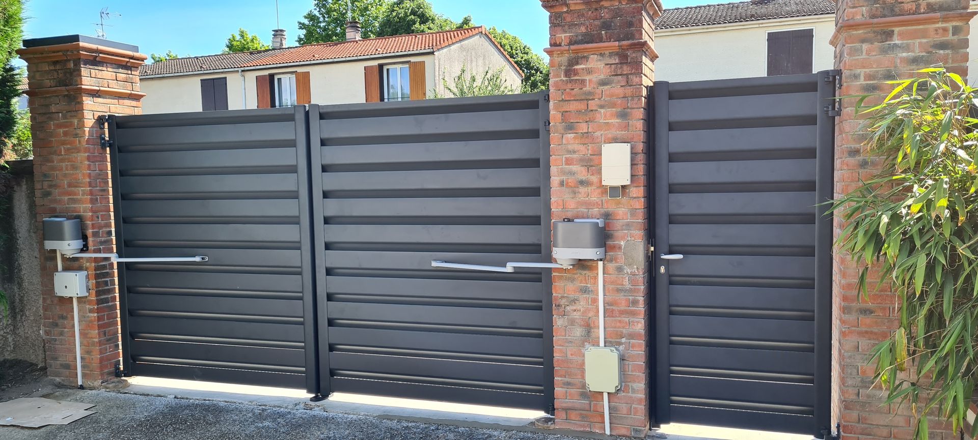 Un portail métallique gris foncé avec des piliers en briques et un système d'ouverture automatique.