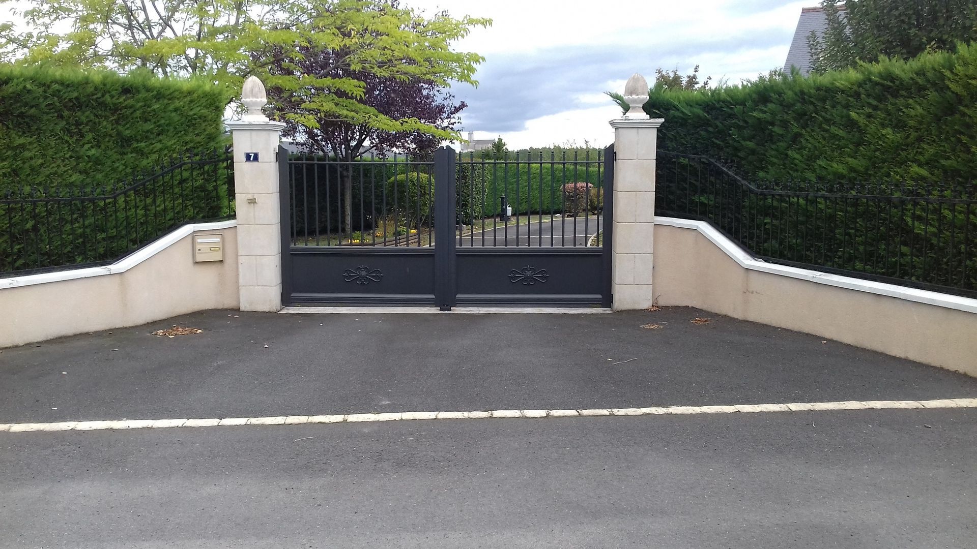 Portail en fer noir dans une allée, flanqué de murs crème, de haies vertes et surmonté de poteaux décoratifs.