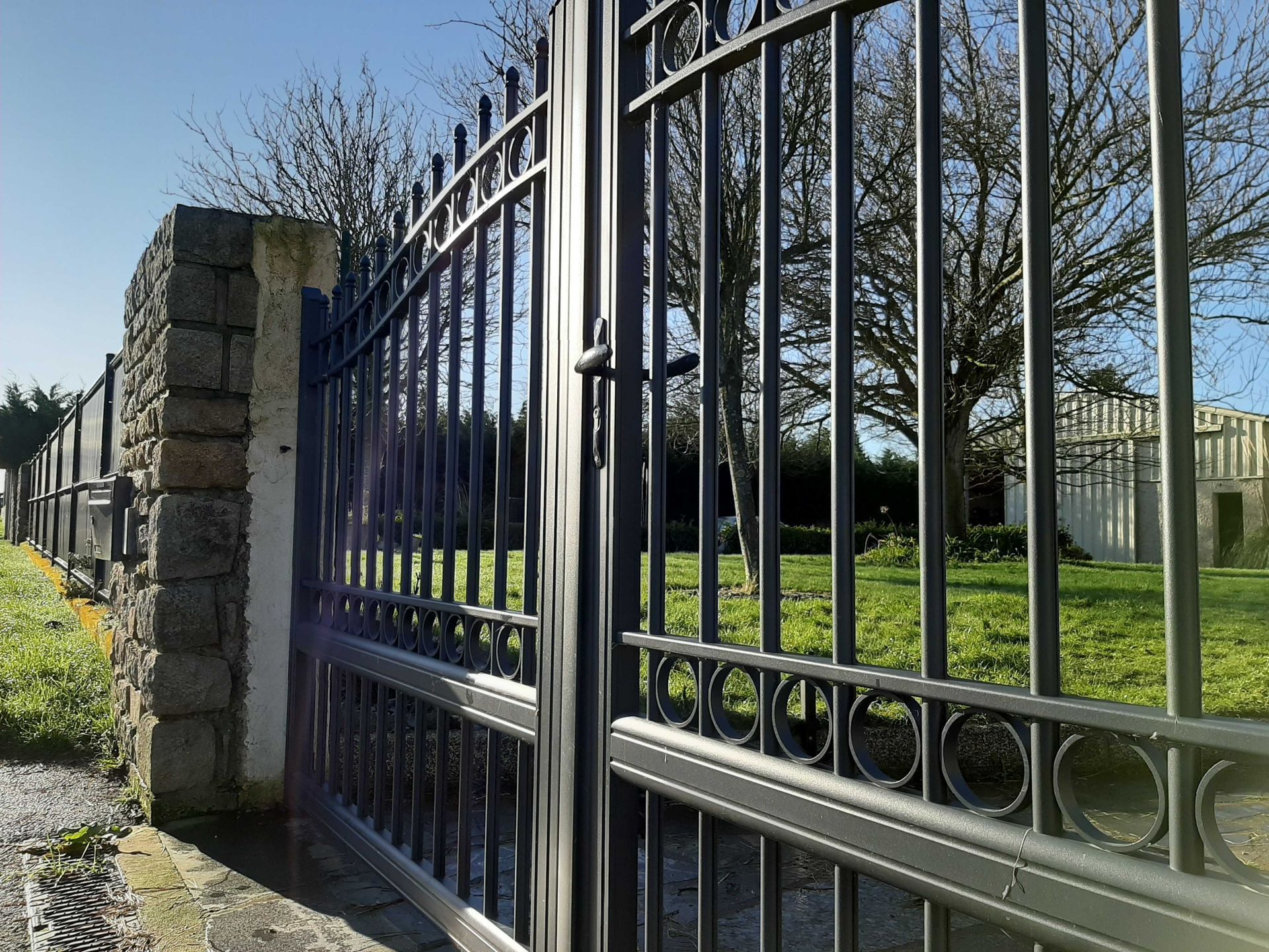 Portail en métal noir et mur de pierre avec de l'herbe verte, des arbres et un ciel ensoleillé en arrière-plan.