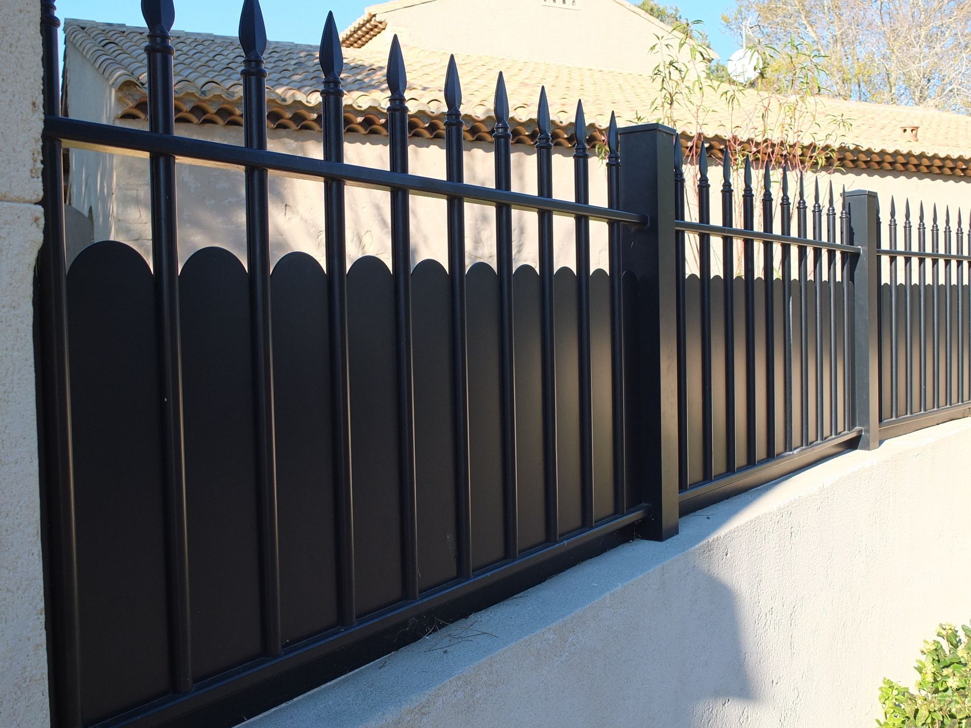 Clôture en métal noir avec panneaux verticaux et sommet en pointe, adossée à un mur blanc, cadre résidentiel.