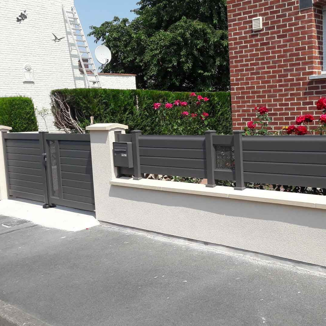 Portail gris et clôture avec poteaux blancs devant une maison en briques avec une haie verte.