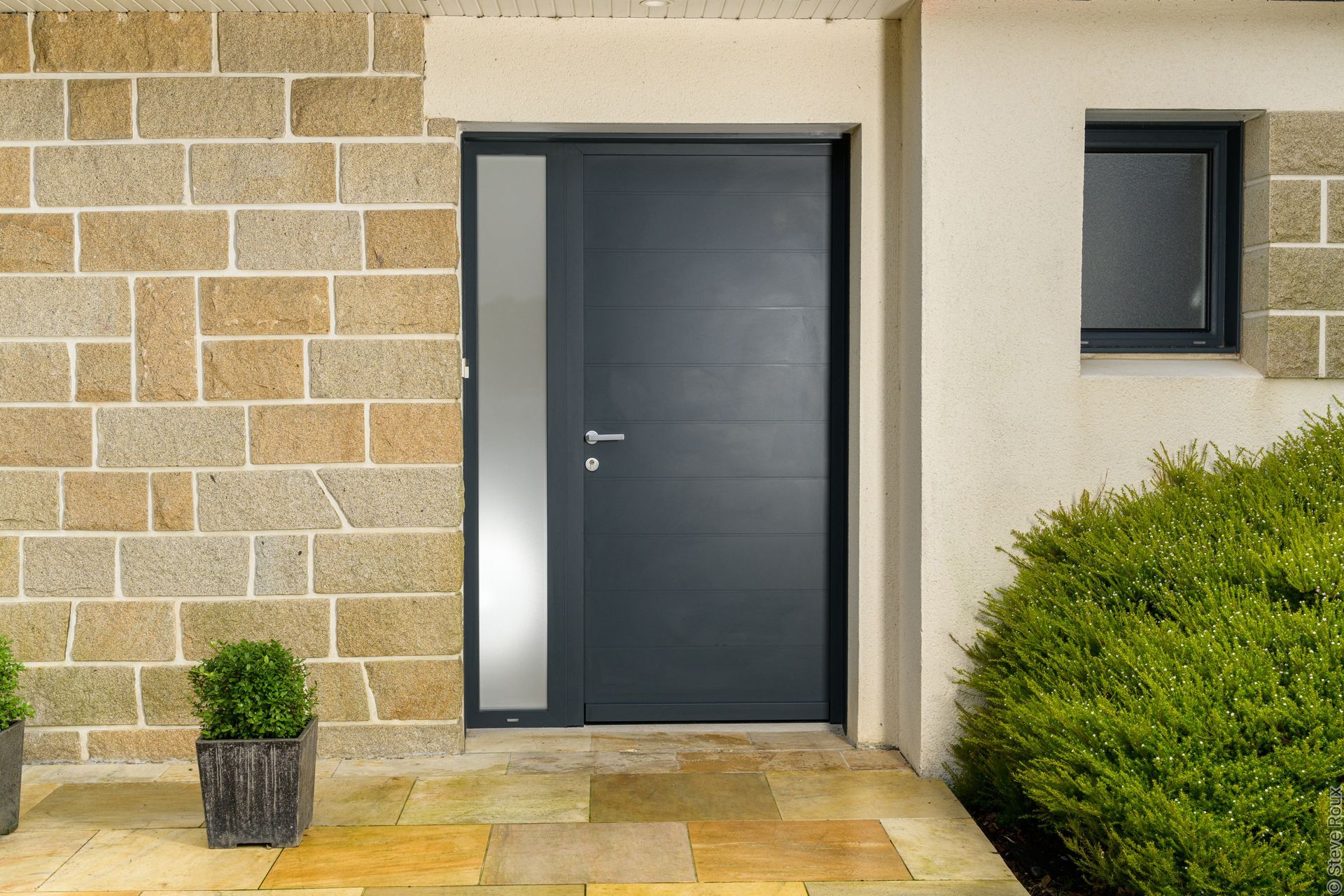 Porte d'entrée gris foncé avec fenêtre latérale, encastrée dans la façade en pierre, avec buisson et plante en pot.