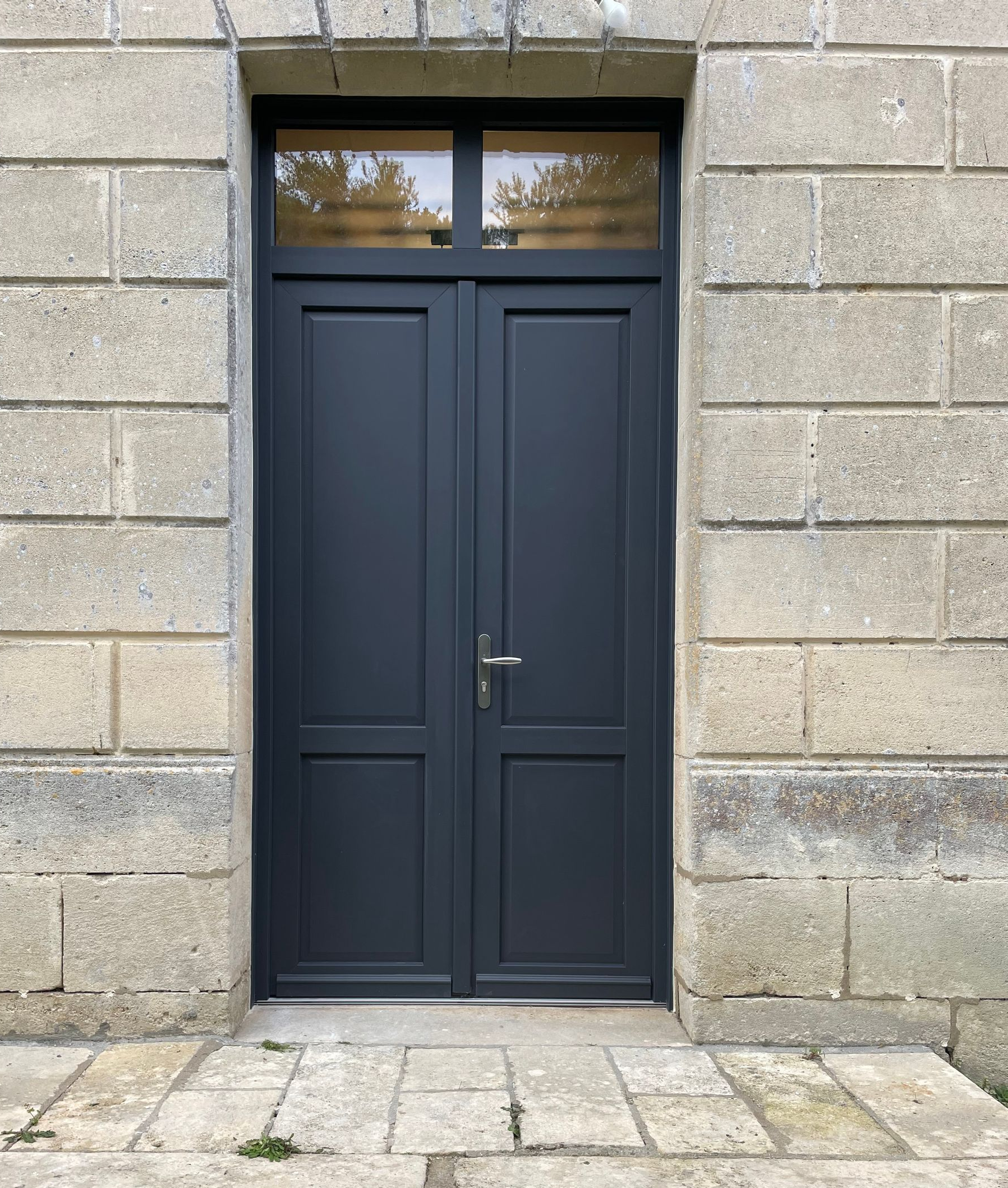 Portes double bleu foncé avec petite fenêtre, encastrées dans un mur en pierre, avec marches en pierre menant vers le haut.