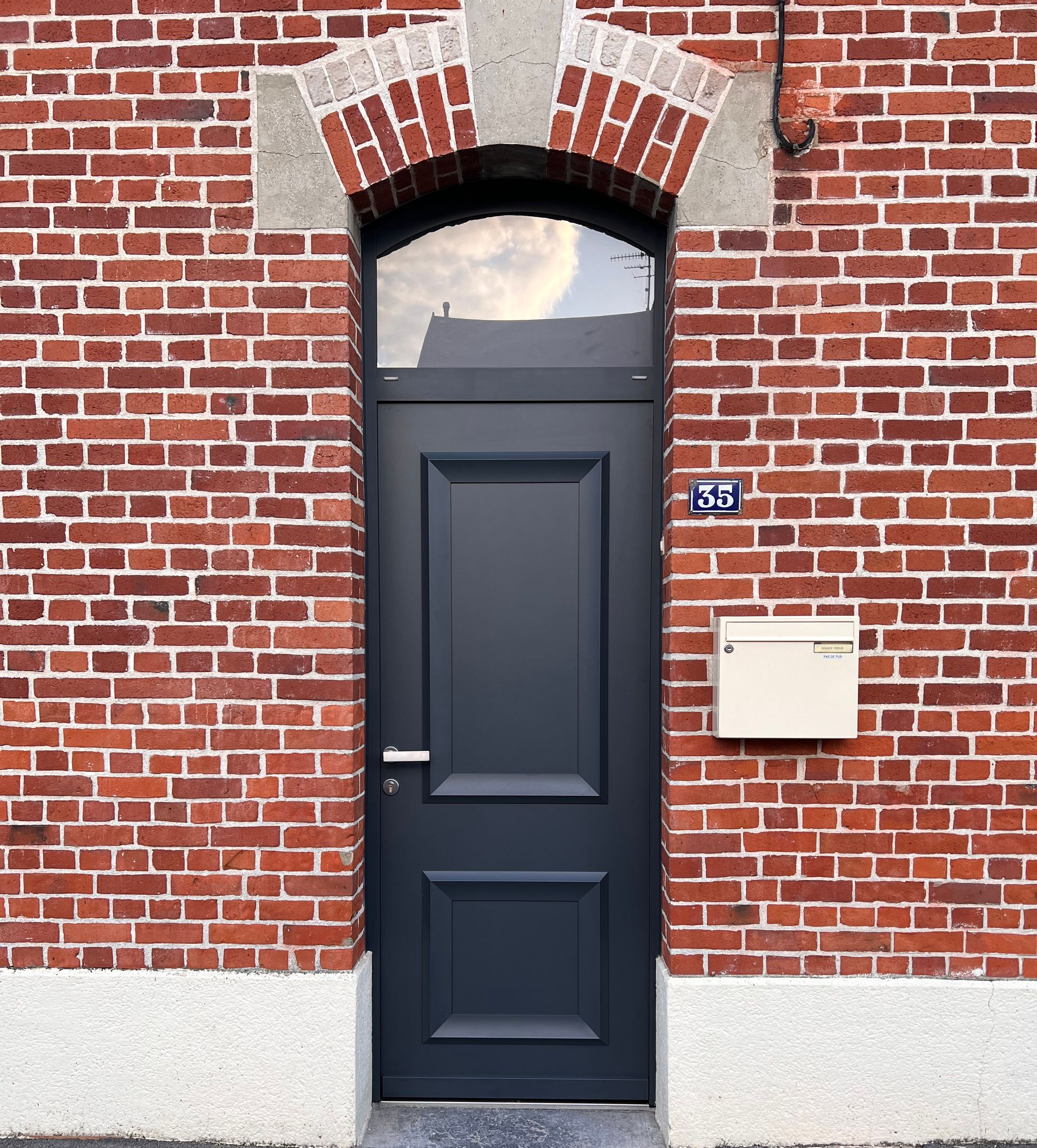 Porte gris foncé avec dessus en verre arqué, encastrée dans un mur de briques rouges.