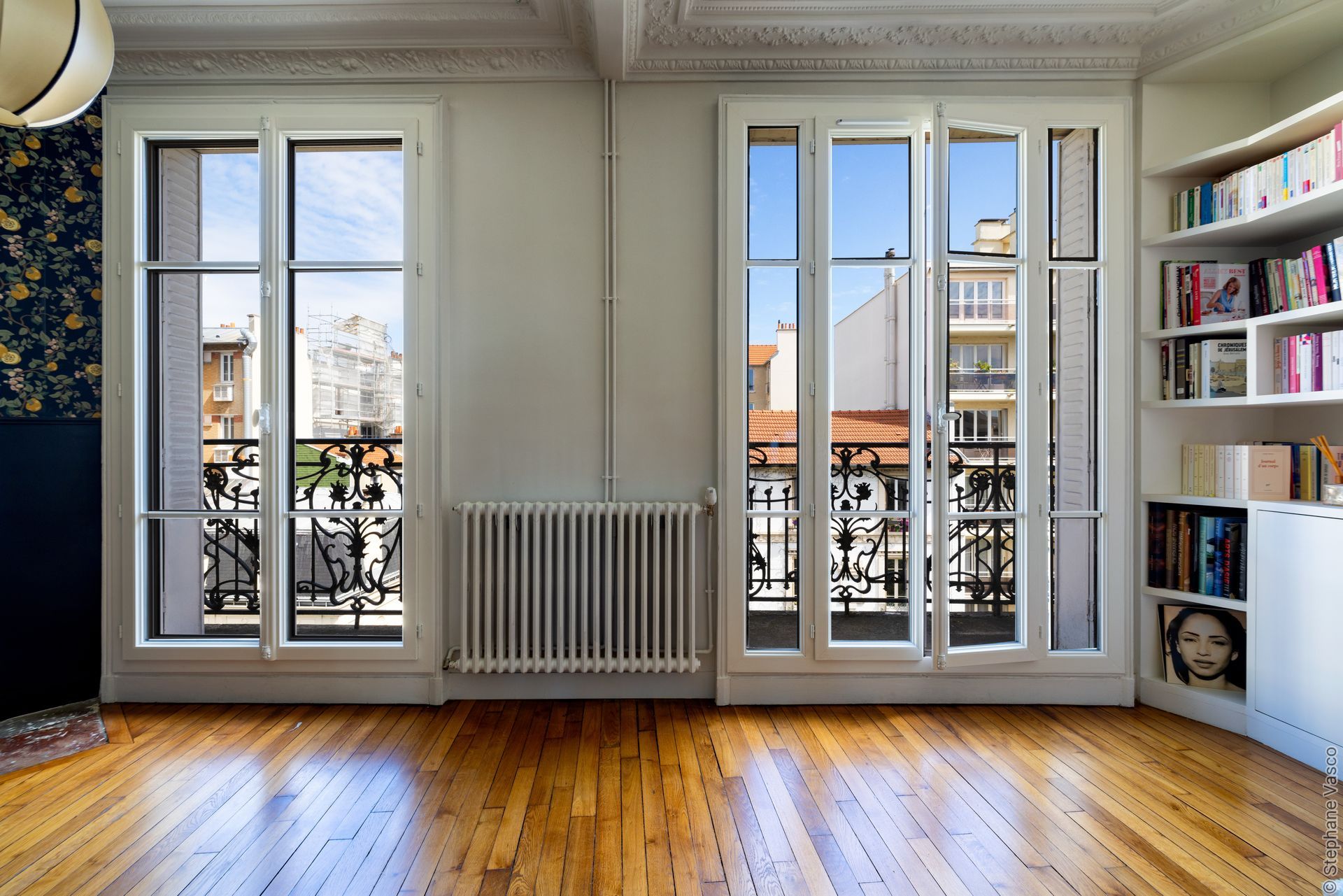 Chambre lumineuse avec deux fenêtres donnant sur une rue de la ville, parquet, radiateur et bibliothèque.