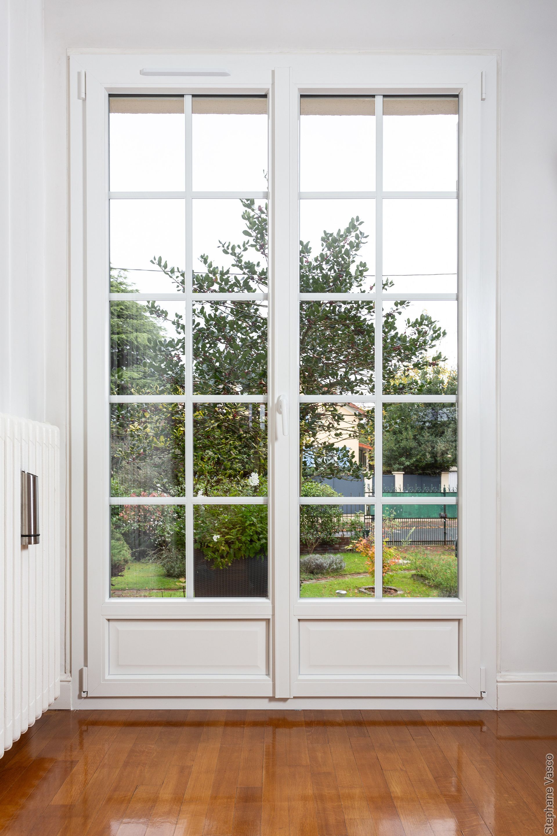 Des portes-fenêtres blanches à plusieurs carreaux s'ouvrent sur un jardin, adossée à un mur blanc et un plancher en bois.