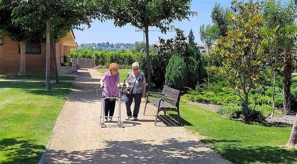 Dues persones caminen per un camí, una amb un caminador. Entorn verd i exuberant amb un banc i arbres.