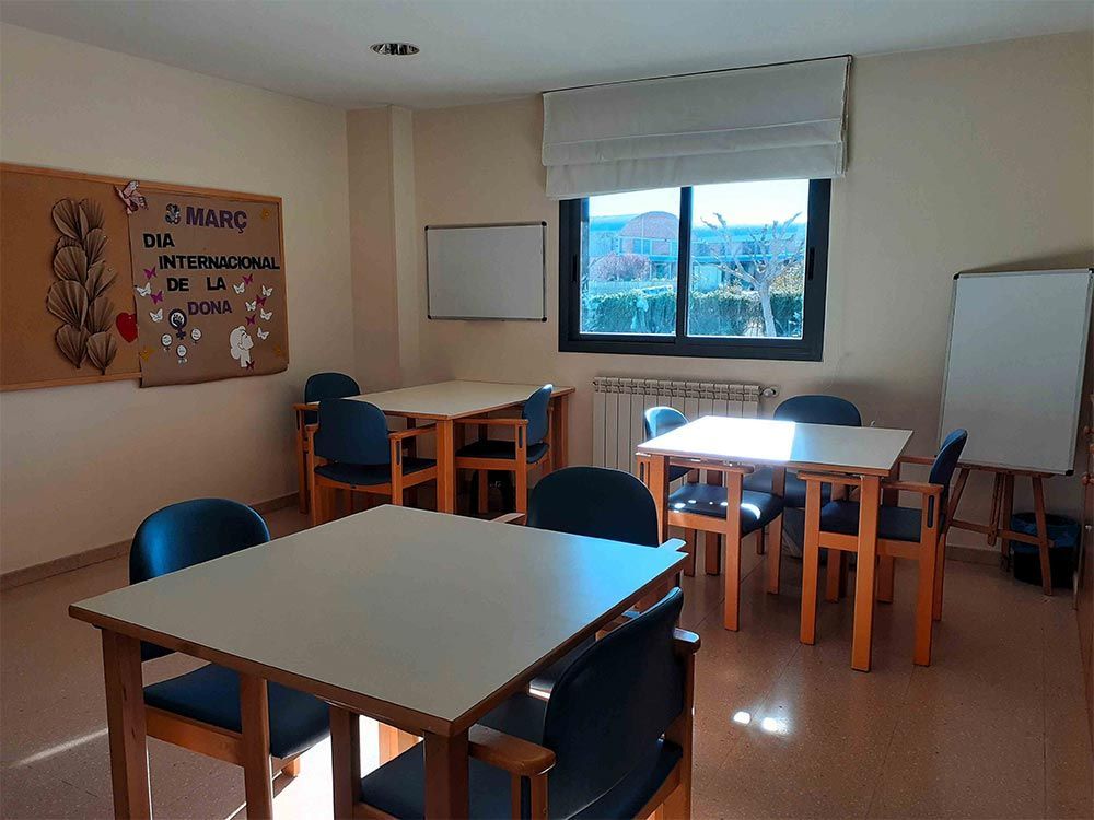 Una sala ben il·luminada amb taules quadrades, cadires, pissarres blanques i una finestra amb vistes a la vegetació.