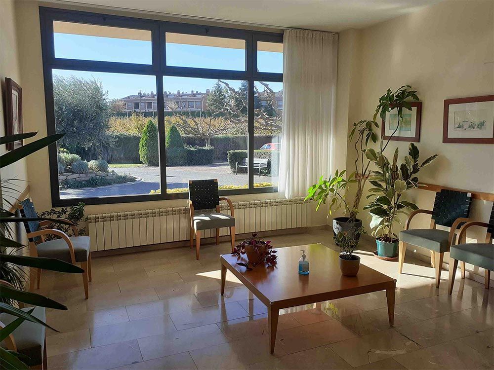 Sala d'espera amb cadires, una taula i plantes al costat d'una gran finestra amb vistes a un jardí.