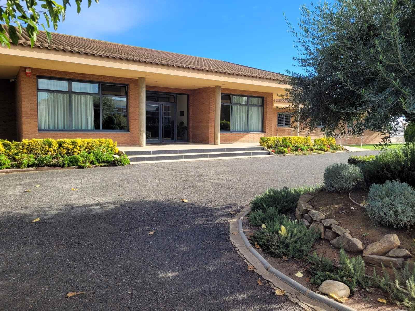 Edifici marró clar amb finestres fosques, entrada i entrada de grava. Arbustos verds i arbres al davant.