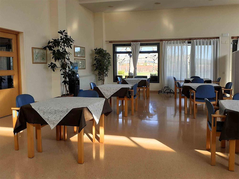 Un menjador lluminós amb taules parades per als àpats, cadires, plantes i finestres amb cortines transparents.