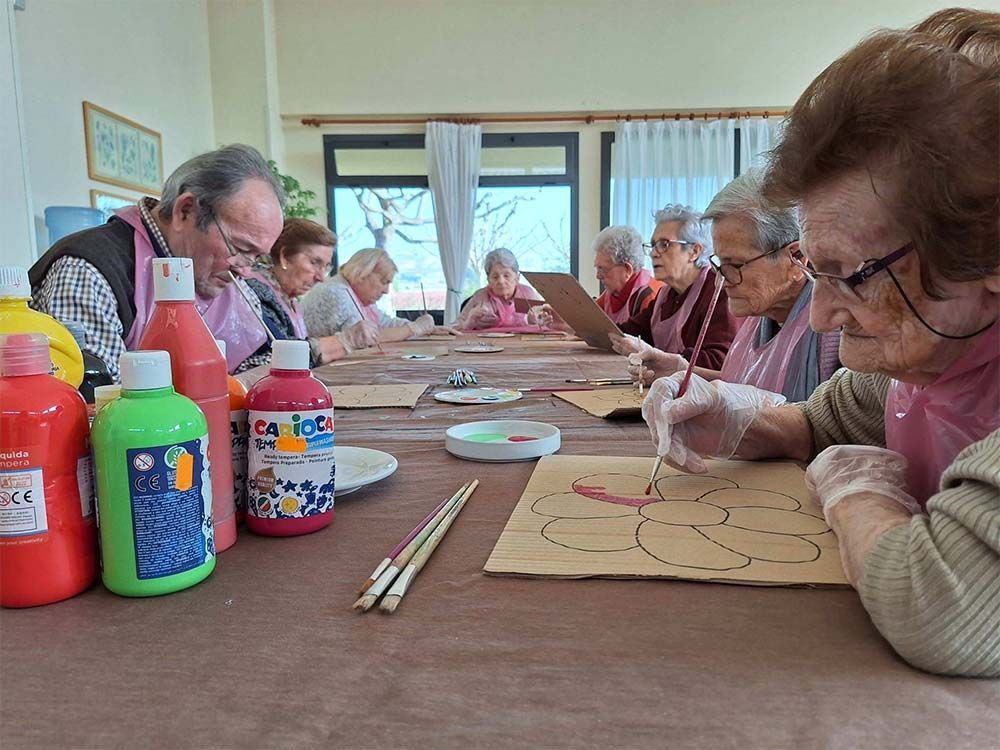 Personas mayores pintando en una mesa, con pinturas y pinceles. Llevan delantales y se concentran en su arte.