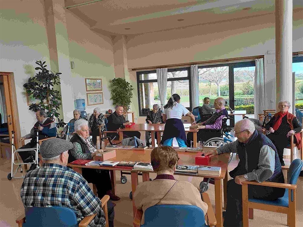 Personas mayores participando en una clase de ejercicio en una sala soleada; el instructor guía los movimientos. Mesas y sillas.