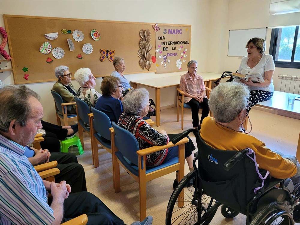 Una mujer habla con un grupo de personas mayores sentadas en una habitación. Hay decoraciones en la pared.
