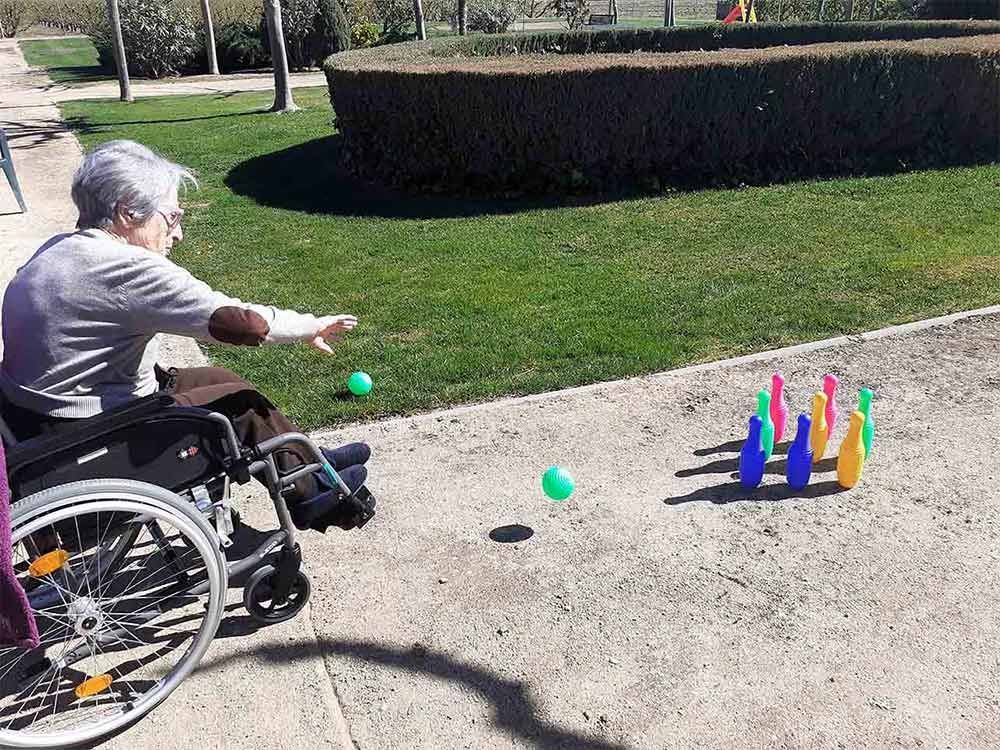 Mujer en silla de ruedas jugando a los bolos al aire libre, apuntando con una bola verde a bolos de colores.