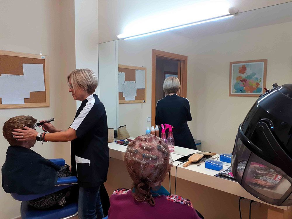 Una dona talla els cabells d'una altra persona en una perruqueria. Una tercera persona seu a prop, amb un tractament al cabell, mirall i estació de perruqueria.
