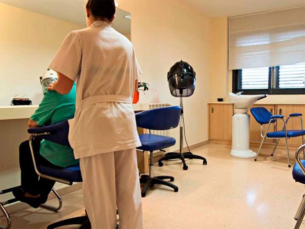 Perruquera pentinant els cabells en una perruqueria. Cadires blaves, campana d'assecat, pica i parets de colors clars.