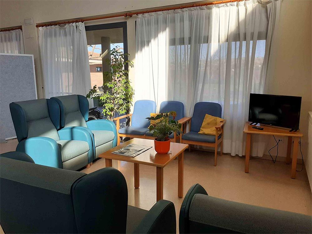 Una sala d'espera amb cadires blaves, una tauleta amb una planta i un televisor a prop d'una finestra amb cortines transparents.