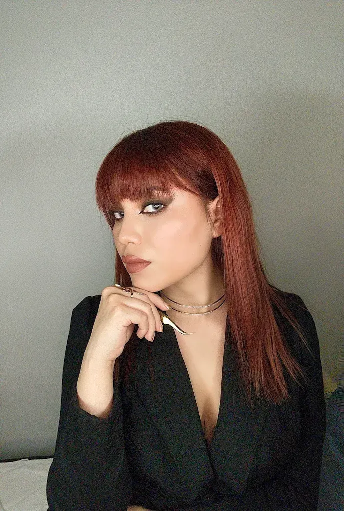 Persona pelirroja con una blusa negra, apoyando la barbilla en la mano sobre un fondo gris liso.