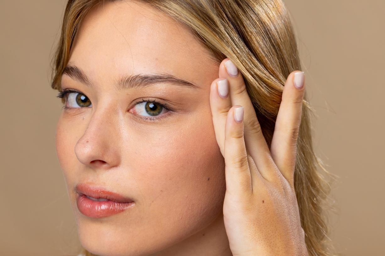 Primer plano de una persona tocándose la sien, mirando a la cámara sobre un fondo beige.